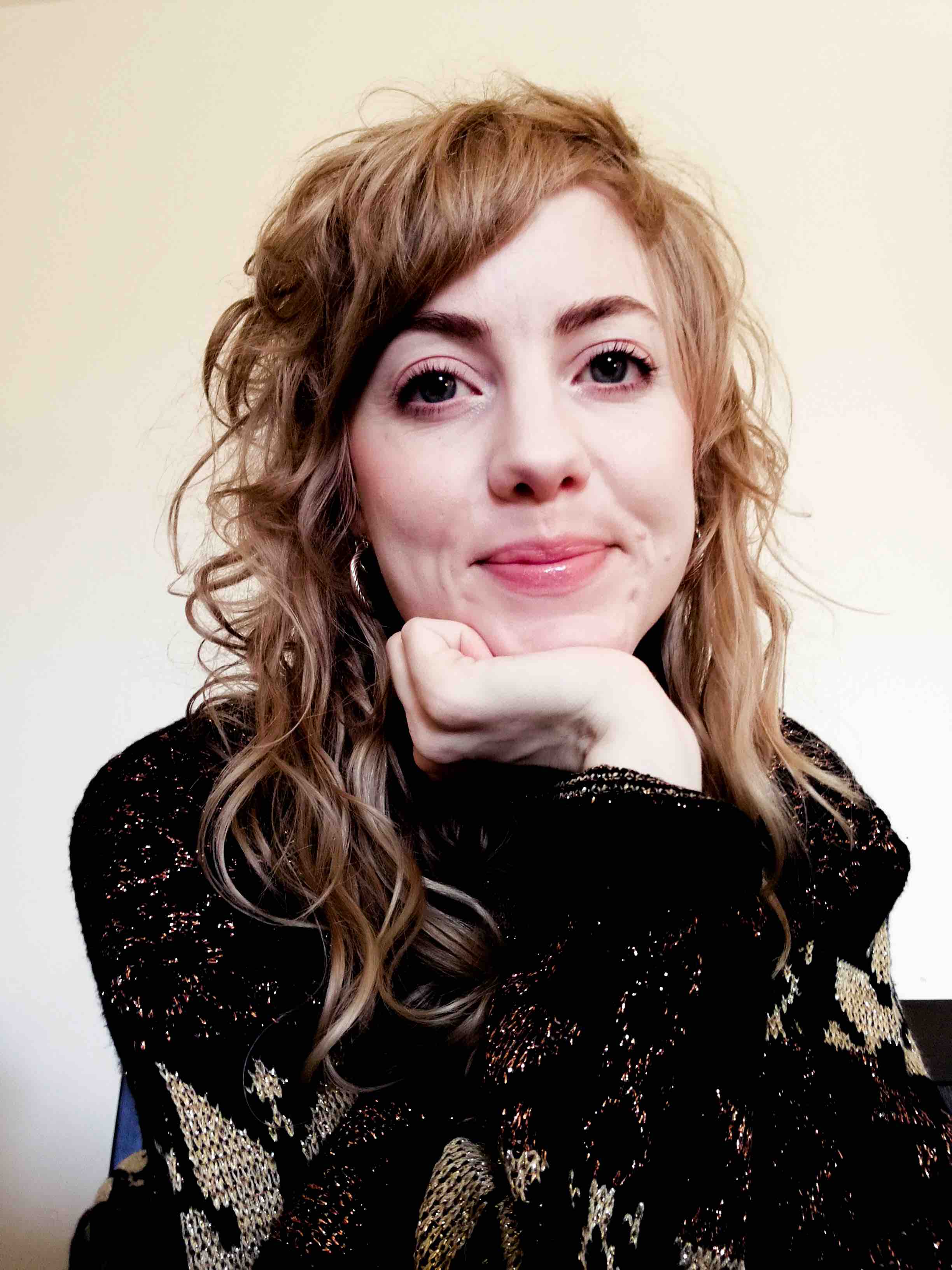 Emma Frid, with blond wavy hair, wearing a black and gold sweater, resting head on hand.